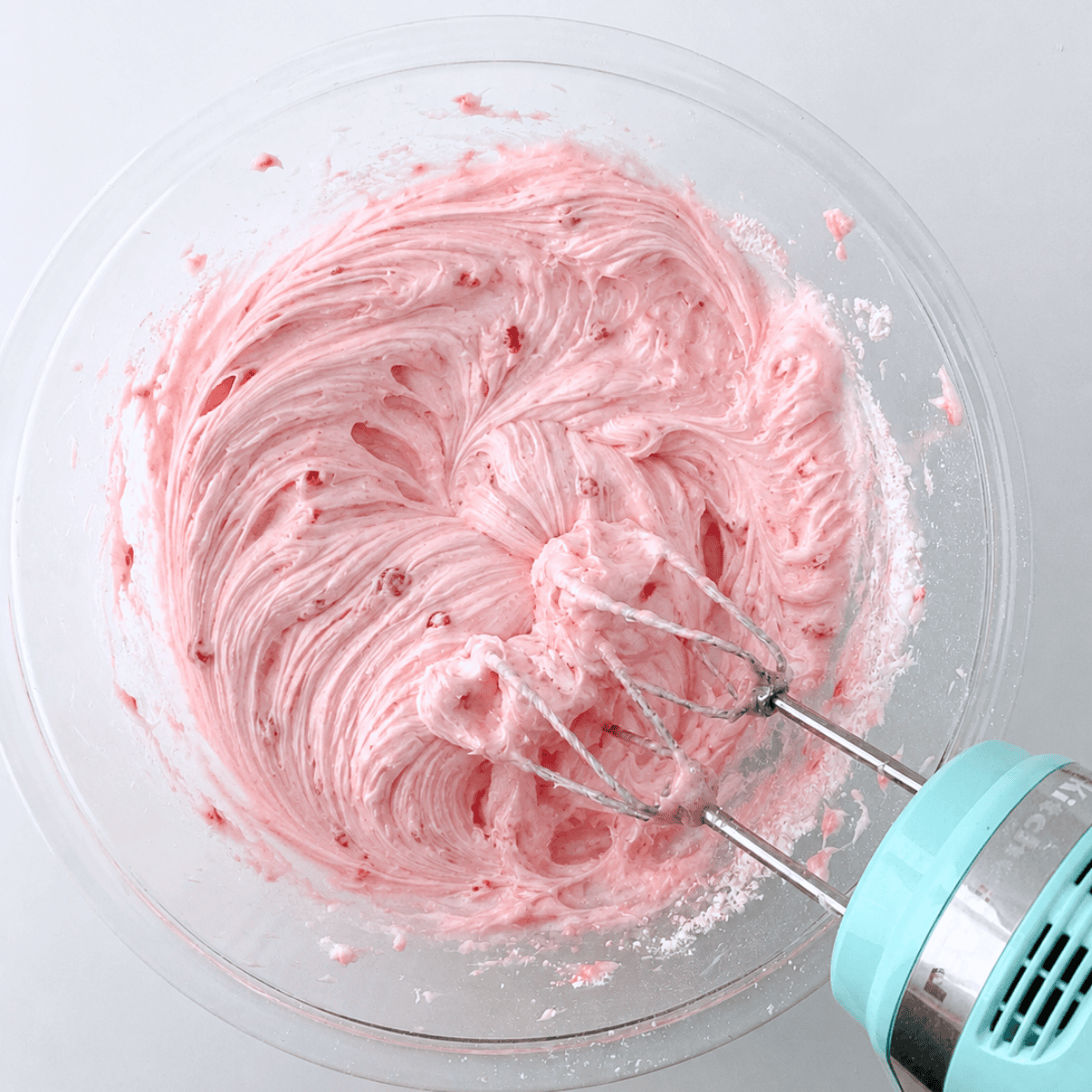 A hand mixer with beaters is mixing pink frosting in a clear glass bowl, creating a smooth, fluffy texture. The background is a plain white surface.
