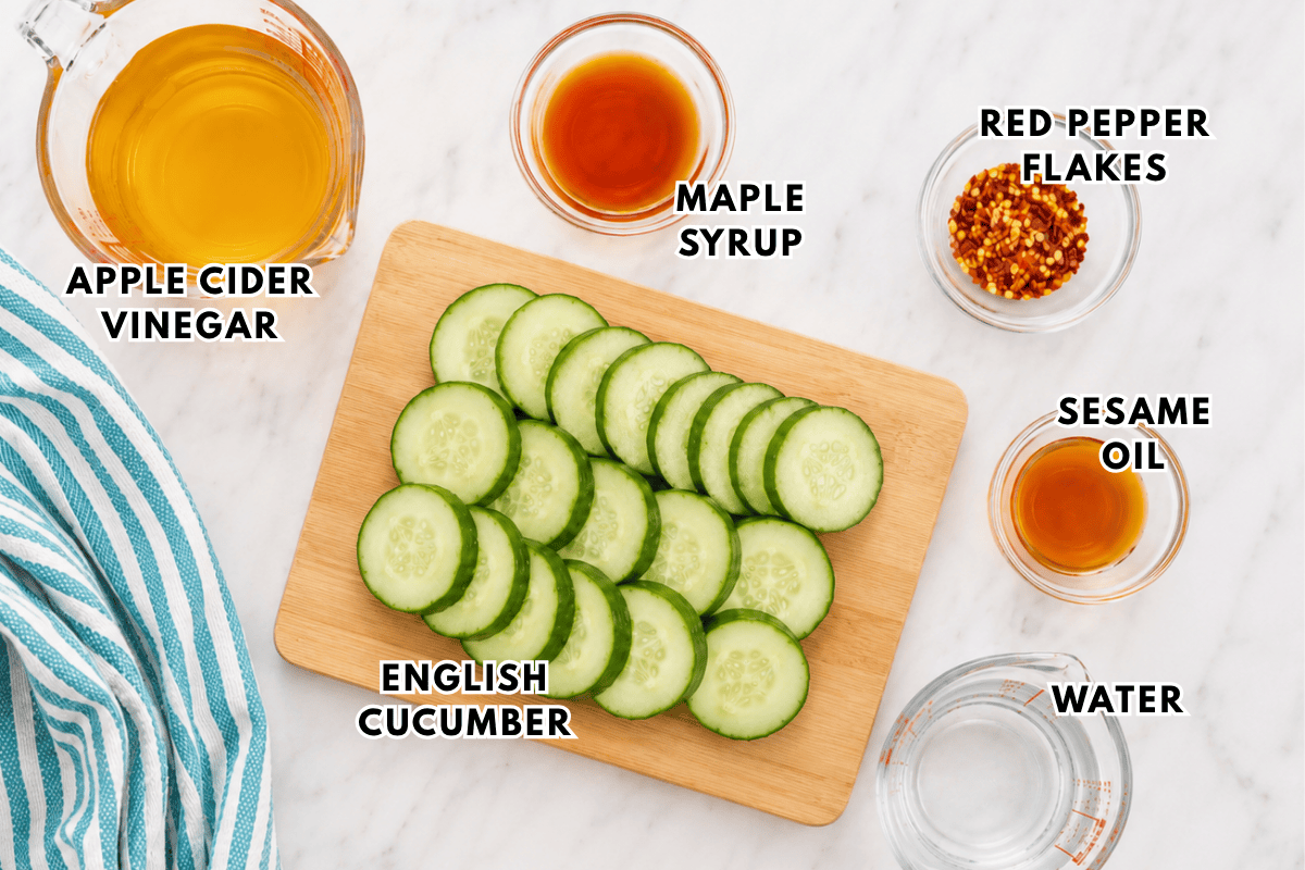 A cutting board with sliced English cucumber is surrounded by bowls of apple cider vinegar, maple syrup, red pepper flakes, sesame oil, and water on a marble surface. Ingredients are labeled.