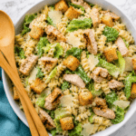 A bowl of Chicken Caesar Pasta Salad with grilled chicken, romaine lettuce, croutons, shaved parmesan, and spiral pasta, served with wooden salad servers on a marble surface.