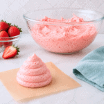 A swirl of pink strawberry frosting sits on parchment paper in the foreground. Behind it is a large glass bowl filled with more frosting, fresh strawberries, a blue towel, and a white tiled wall.