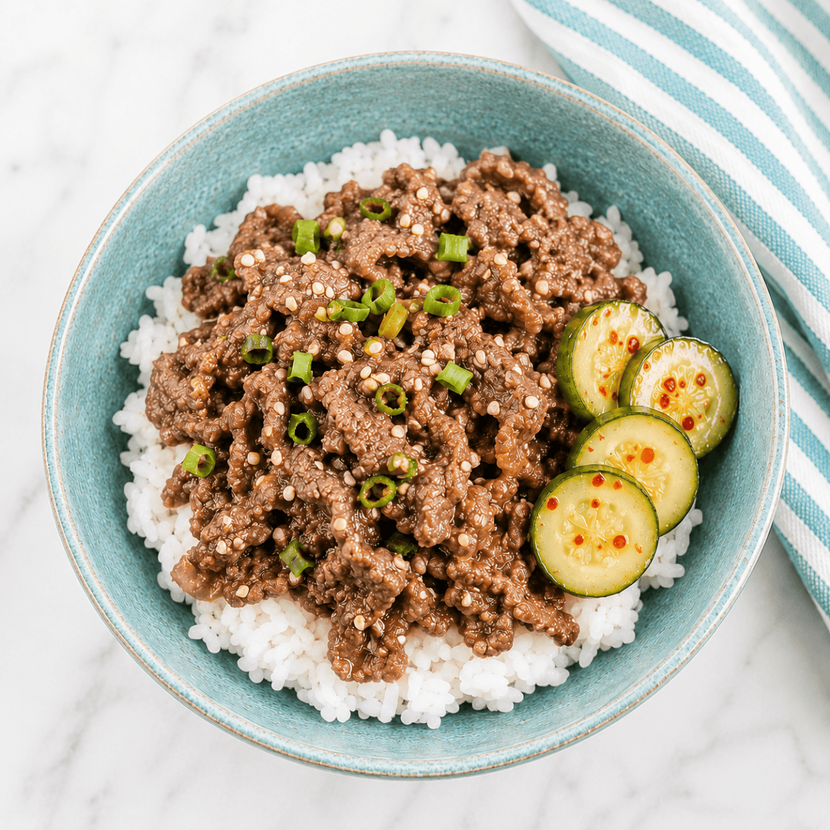 A blue bowl filled with white rice topped with seasoned ground beef, sliced green onions, sesame seeds, and sliced cucumbers. A striped cloth napkin is partially visible beside the bowl.