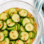 A glass bowl filled with sliced cucumbers in a light dressing, garnished with red pepper flakes. A striped cloth napkin is partially visible beside the bowl on a marble surface.