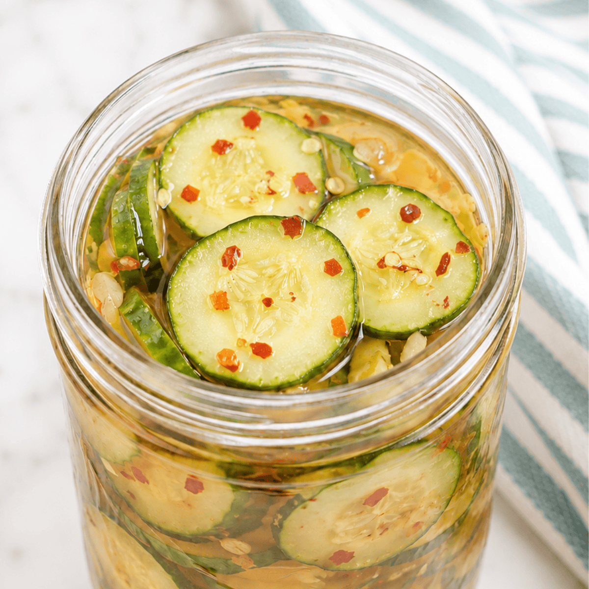 A glass jar filled with sliced cucumbers in a pickling liquid, topped with red pepper flakes and seeds, sits on a marble surface next to a white and teal striped cloth.