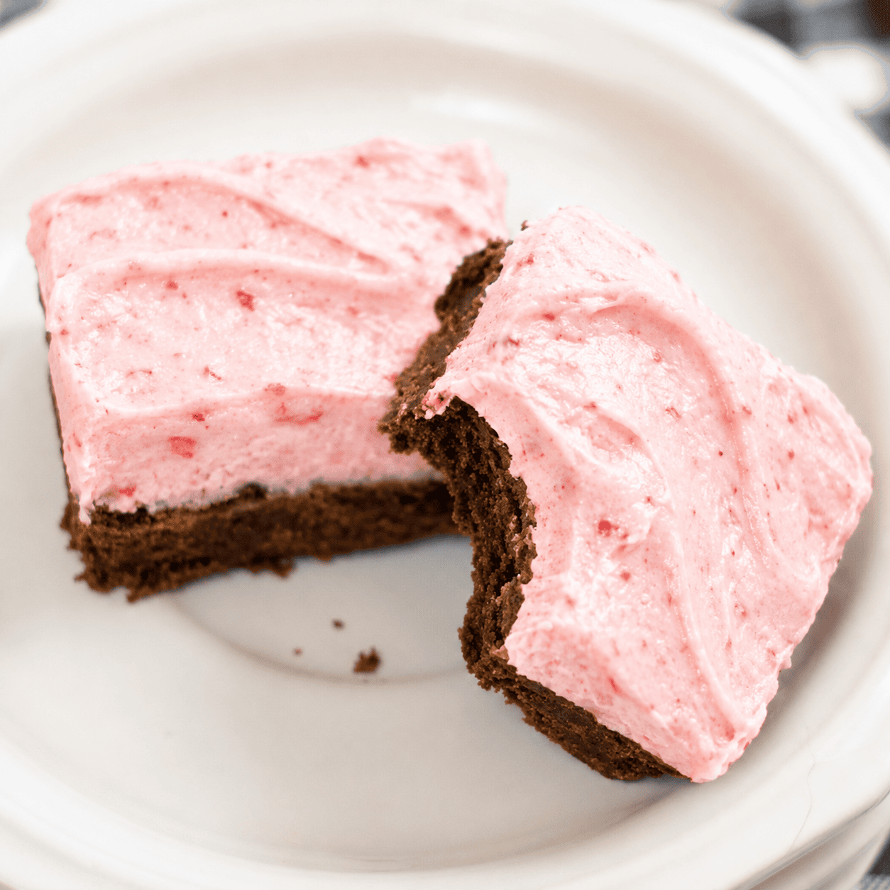 Two chocolate cake squares with fresh strawberry frosting on a white plate, one with a bite taken out of it.
