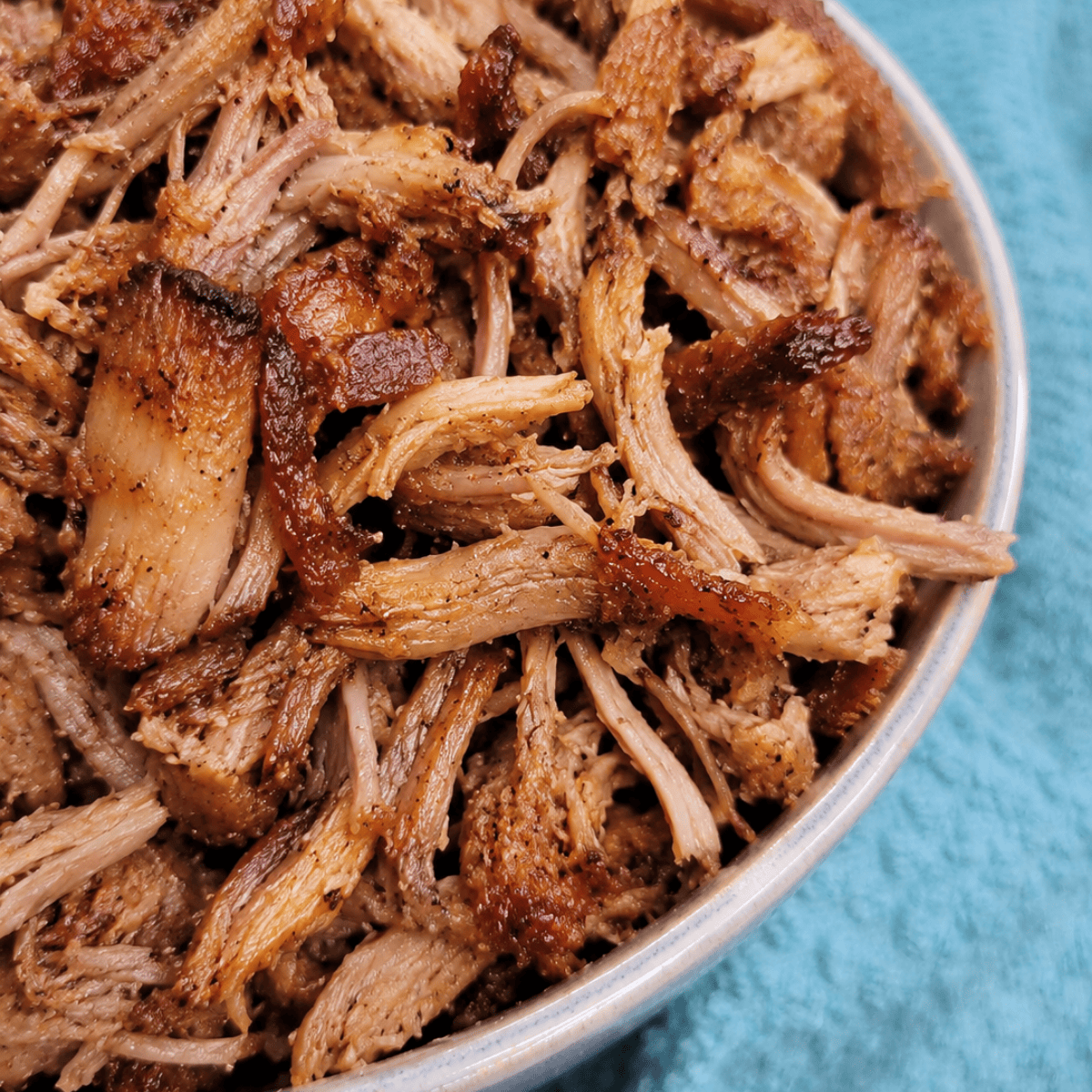 A close-up of a bowl filled with shredded, seasoned, and crispy cooked pork, with tender and browned edges, resting on a blue textured cloth.