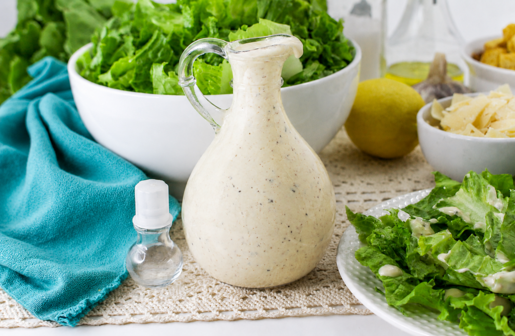 A glass pitcher filled with creamy Caesar salad dressing sits on a table, surrounded by bowls of fresh romaine lettuce, parmesan cheese, croutons, a lemon, and a Chicken Caesar Pasta Salad ready to be enjoyed.