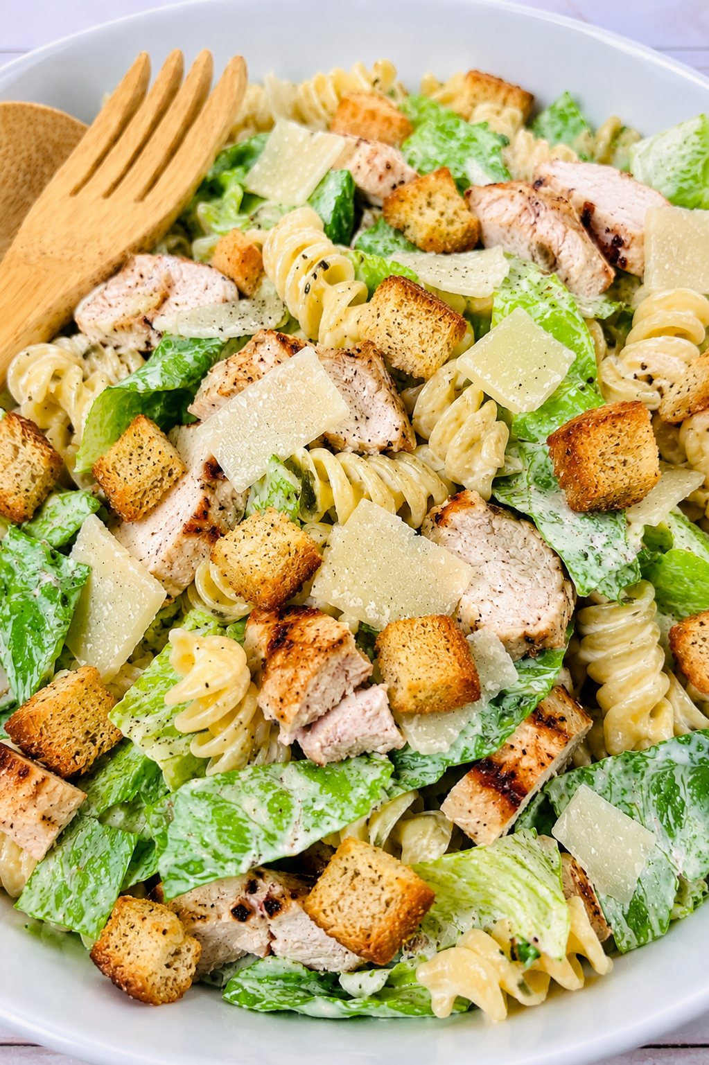 A bowl of Caesar pasta salad with grilled chicken, rotini pasta, romaine lettuce, croutons, and shaved parmesan cheese, served with wooden salad tongs.