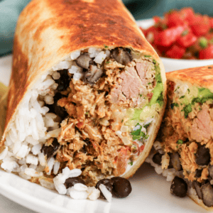 A close-up of a burrito cut in half, showing rice, black beans, shredded meat, guacamole, and salsa filling, with a crispy tortilla and salsa in the background.