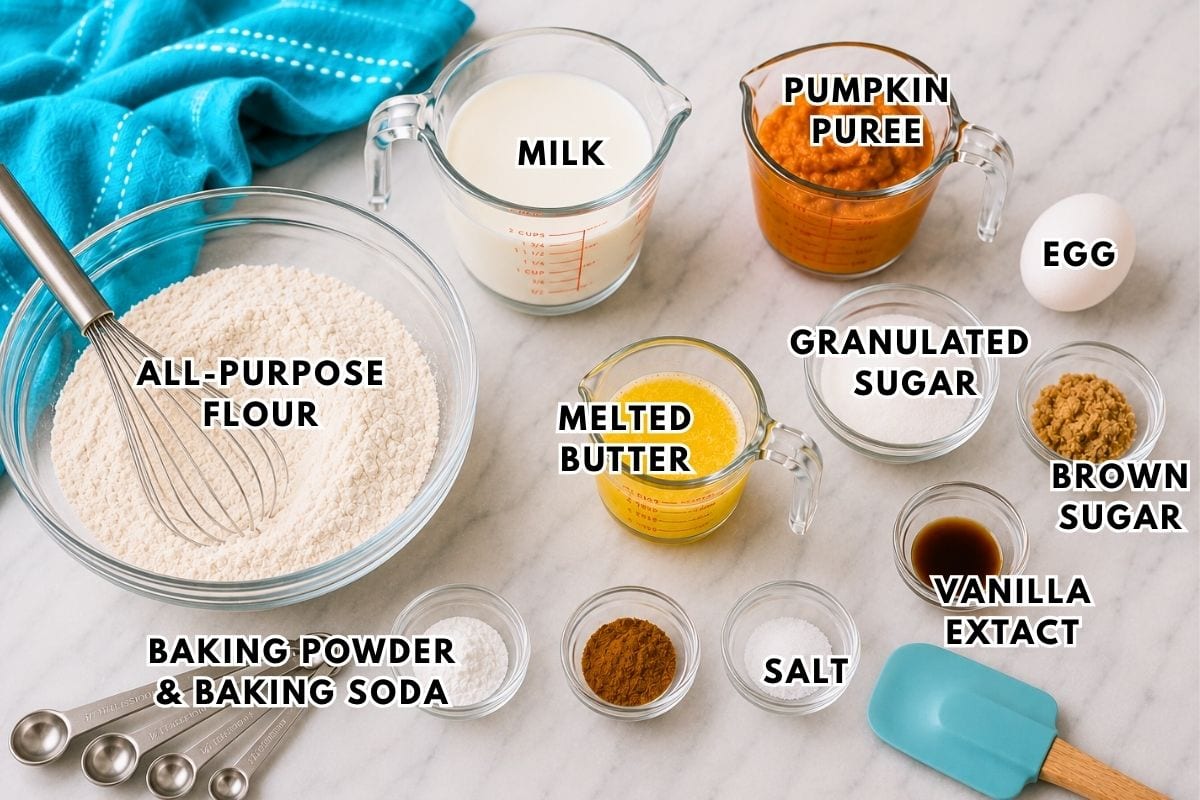 Baking ingredients on a counter: flour, milk, pumpkin puree, egg, granulated sugar, brown sugar, melted butter, baking powder, baking soda, salt, vanilla extract, and a whisk with blue measuring spoons.