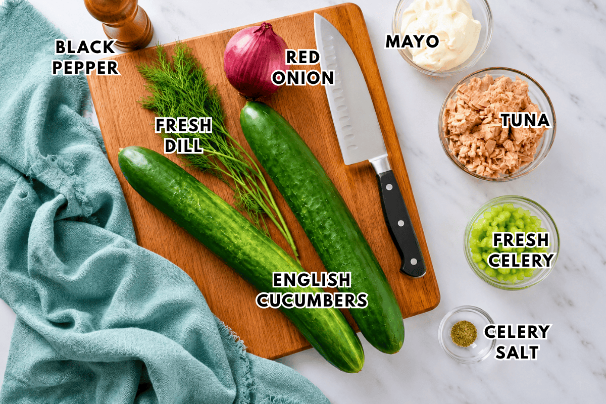 Ingredients for tuna salad cucumbers on a marble counter: black pepper, red onion, mayo, canned tuna, fresh celery, celery salt, fresh dill, and two English cucumbers on a wooden board with a knife.