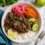 Korean Beef and Rice: A bowl with shredded beef, white rice, sliced cucumbers, pickled red onions, shredded carrots, a lime wedge, and sesame seeds, garnished with green onions and black pepper, on a marble surface with a fork and cilantro nearby.