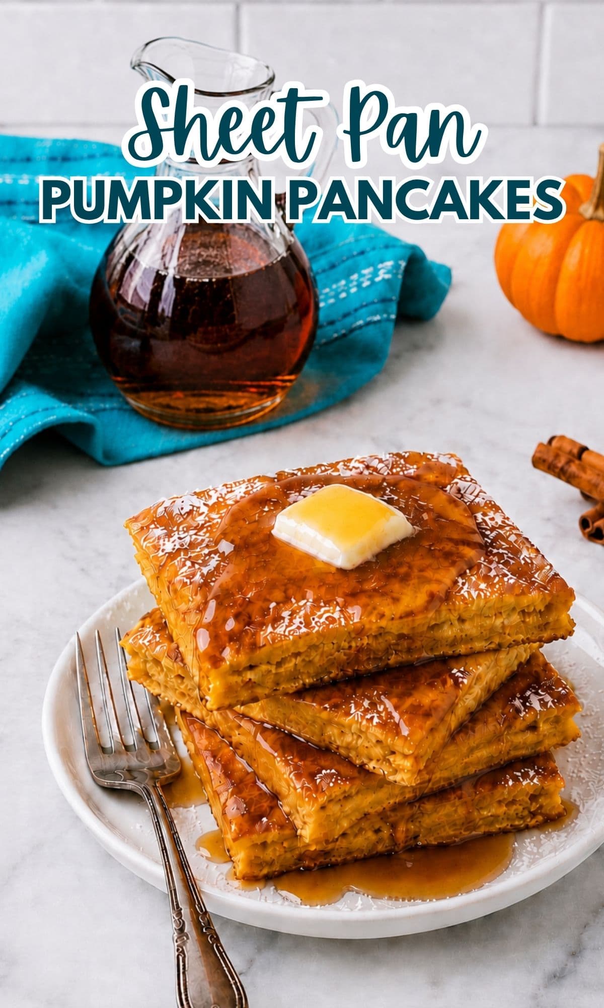A stack of square pumpkin pancakes topped with butter and syrup sits on a white plate with two forks. A glass jar of syrup, a blue cloth, a pumpkin, and cinnamon sticks are in the background.
