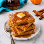 A stack of square pancakes topped with butter and syrup sits on a plate with a fork. Cinnamon sticks, a pumpkin, and a syrup bottle are in the background on a marble surface.