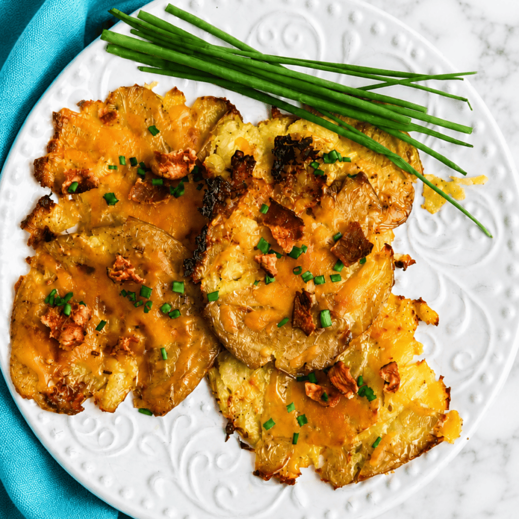 A white plate with smashed roasted potatoes topped with melted cheese, bacon bits, and chopped chives, garnished with whole fresh chives on the side. The plate sits on a marble surface with a blue napkin.