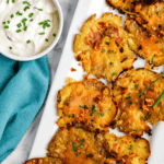 Crispy smashed potatoes topped with melted cheese and chopped chives on a white tray, served with a bowl of sour cream garnished with chives, next to a teal napkin.