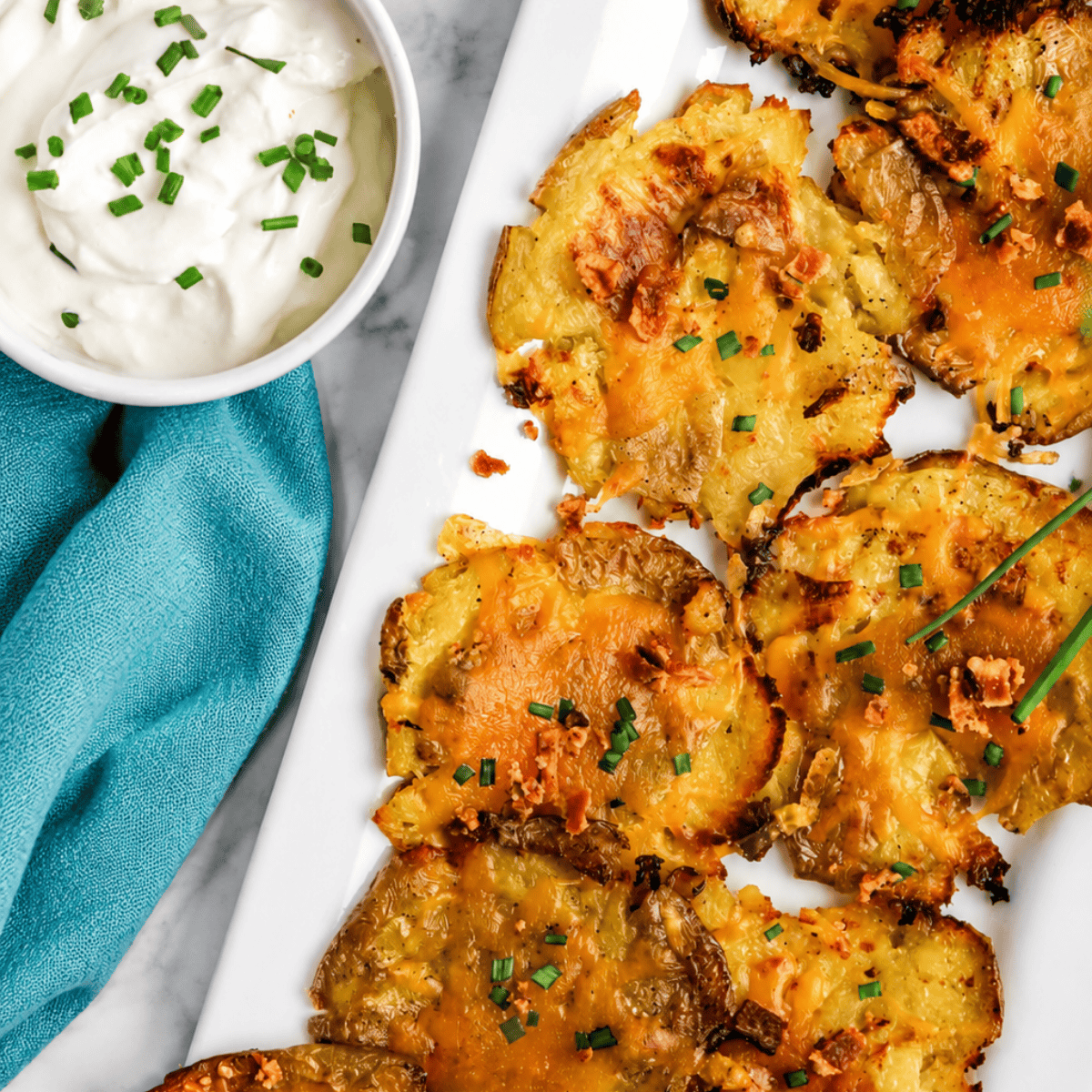 Crispy smashed potatoes topped with melted cheese and chopped chives on a white tray, served with a bowl of sour cream garnished with chives, next to a teal napkin.
