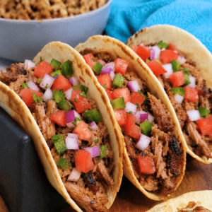 Three soft tacos filled with shredded meat, diced tomatoes, red onions, and green peppers are lined up on a holder, with a bowl of extra meat and a blue cloth in the background.