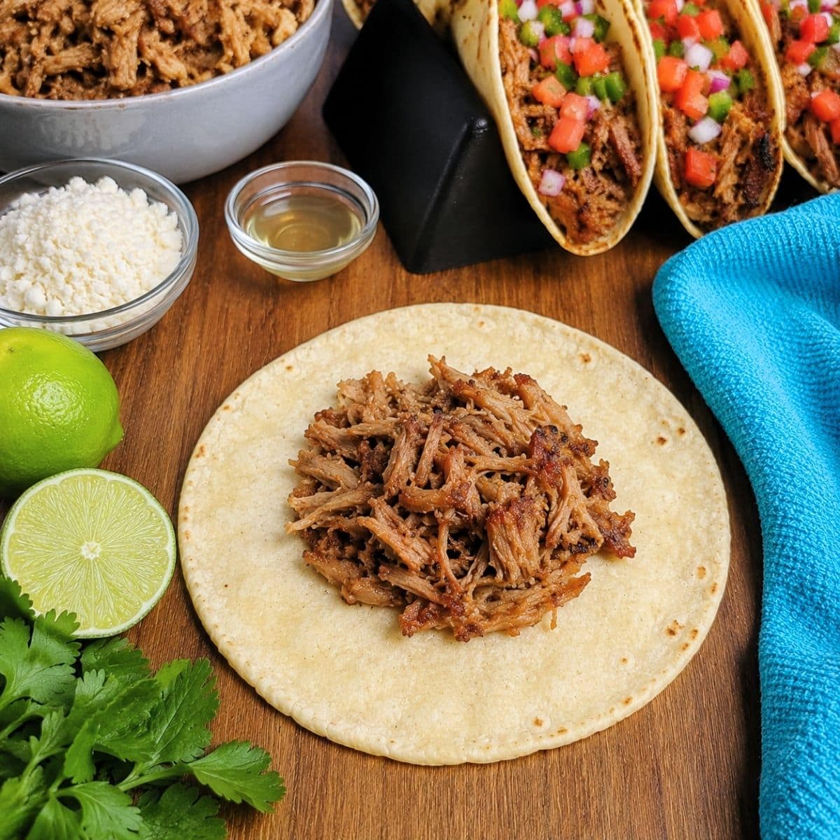 Savory shredded pork sits on a tortilla surrounded by limes, cilantro, cheese, a bowl of Pork Carnitas Tacos filling, a small glass bowl, three filled tacos, and a blue towel on a wooden surface.