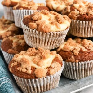 A close-up of several Pumpkin Cream Cheese Muffins, crowned with a buttery streusel topping and nestled in white paper liners, stacked together on a metal tray next to a blue striped cloth.