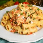 A white plate filled with rigatoni pasta mixed with crumbled cheese, roasted tomatoes, and chopped fresh basil. The pasta is coated in a creamy, slightly orange-colored sauce.