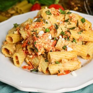A white plate filled with rigatoni pasta mixed with crumbled cheese, roasted tomatoes, and chopped fresh basil. The pasta is coated in a creamy, slightly orange-colored sauce.
