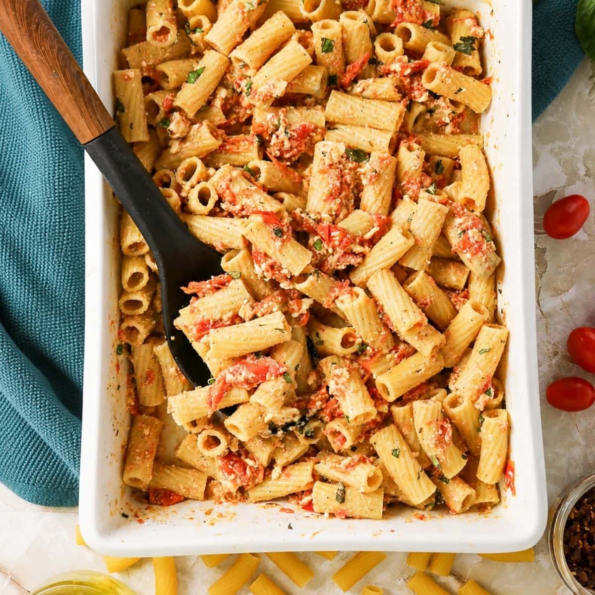 Baked rigatoni pasta mixed with creamy sauce, cherry tomatoes, and herbs in a white casserole dish, with a black serving spoon. Uncooked pasta, tomatoes, and a green towel are nearby.