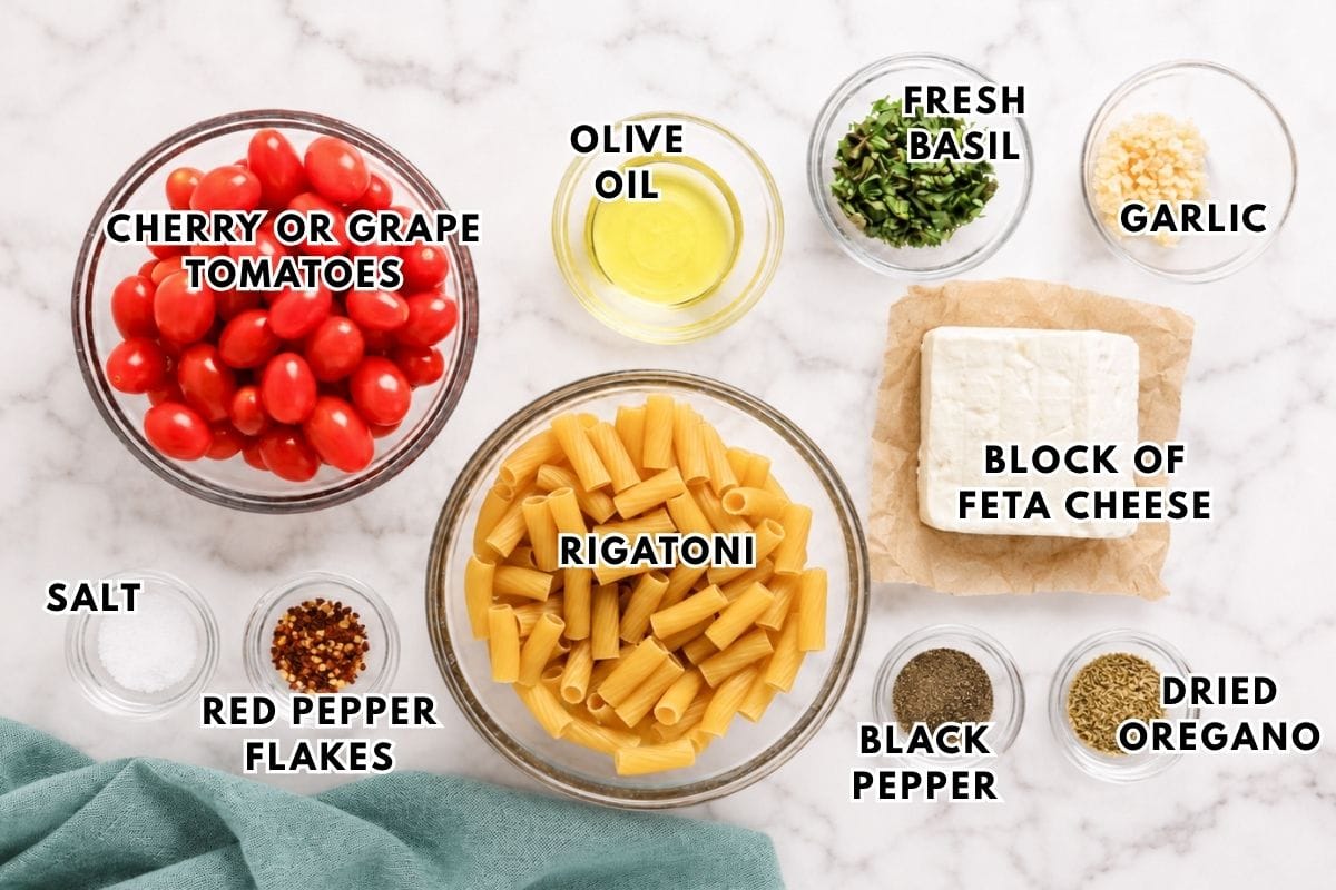 Ingredients for a pasta dish: cherry tomatoes, olive oil, fresh basil, garlic, feta cheese, rigatoni, salt, red pepper flakes, black pepper, and dried oregano, all arranged in bowls on a marble surface.