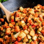 A close-up of a cast iron skillet filled with diced, cooked vegetables including potatoes, sweet potatoes, bell peppers, and herbs, with a wooden spoon stirring the mixture.