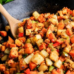 A close-up of a cast iron skillet filled with diced, cooked vegetables including potatoes, sweet potatoes, bell peppers, and herbs, with a wooden spoon stirring the mixture.