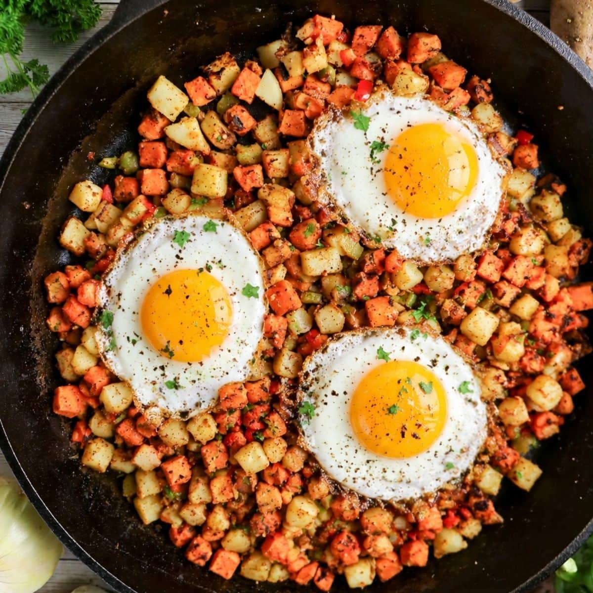 A skillet filled with three sunny-side-up eggs on top of a mix of diced potatoes and sweet potatoes, garnished with herbs.