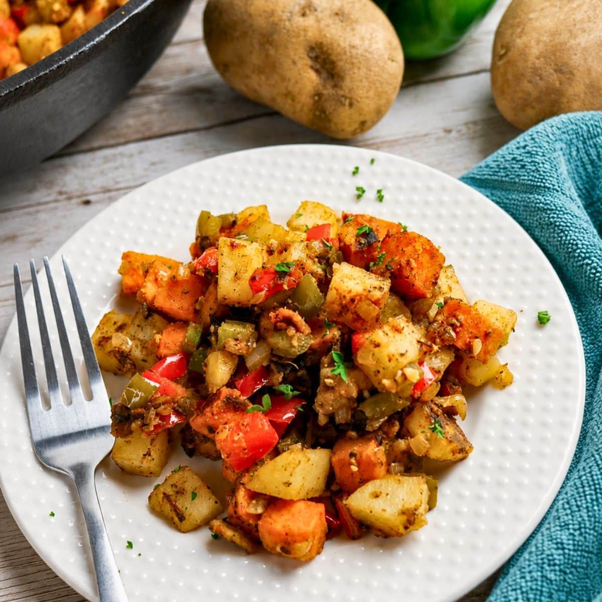 A white plate with a colorful vegetable hash, including potatoes, carrots, and peppers, garnished with herbs. A fork is on the plate, and whole potatoes and a blue towel are in the background.