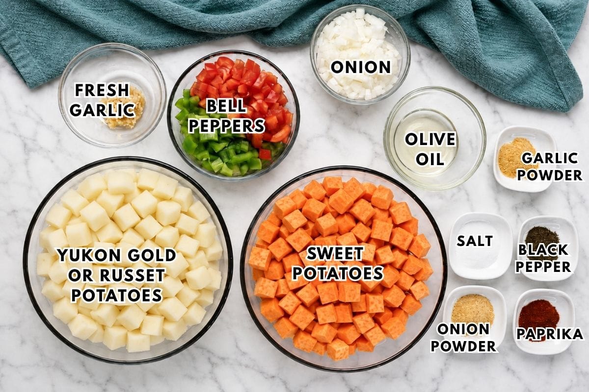 Bowls of cubed Yukon gold, russet, and sweet potatoes, diced bell peppers, onions, and fresh garlic, with small dishes of olive oil, salt, pepper, paprika, garlic powder, and onion powder, on a marble surface.