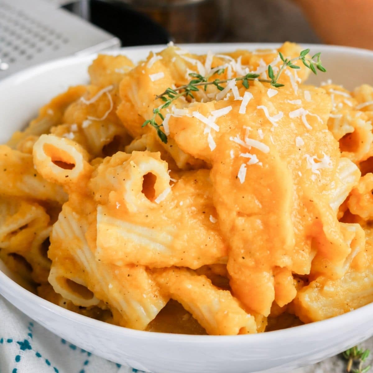 A bowl of rigatoni pasta coated in a creamy orange sauce, topped with grated cheese and a sprig of fresh thyme.