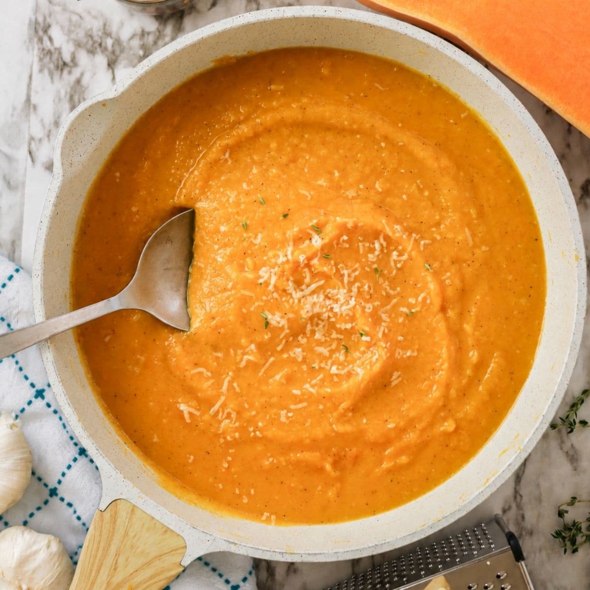 A bowl of creamy orange soup topped with shredded cheese, with a spoon resting inside. Garlic, cheese, and a grater are nearby on a marble surface.