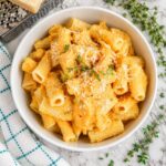 A bowl of rigatoni pasta in a creamy orange sauce, topped with grated cheese and fresh thyme, sits on a marble surface next to a checkered towel and sprigs of thyme.