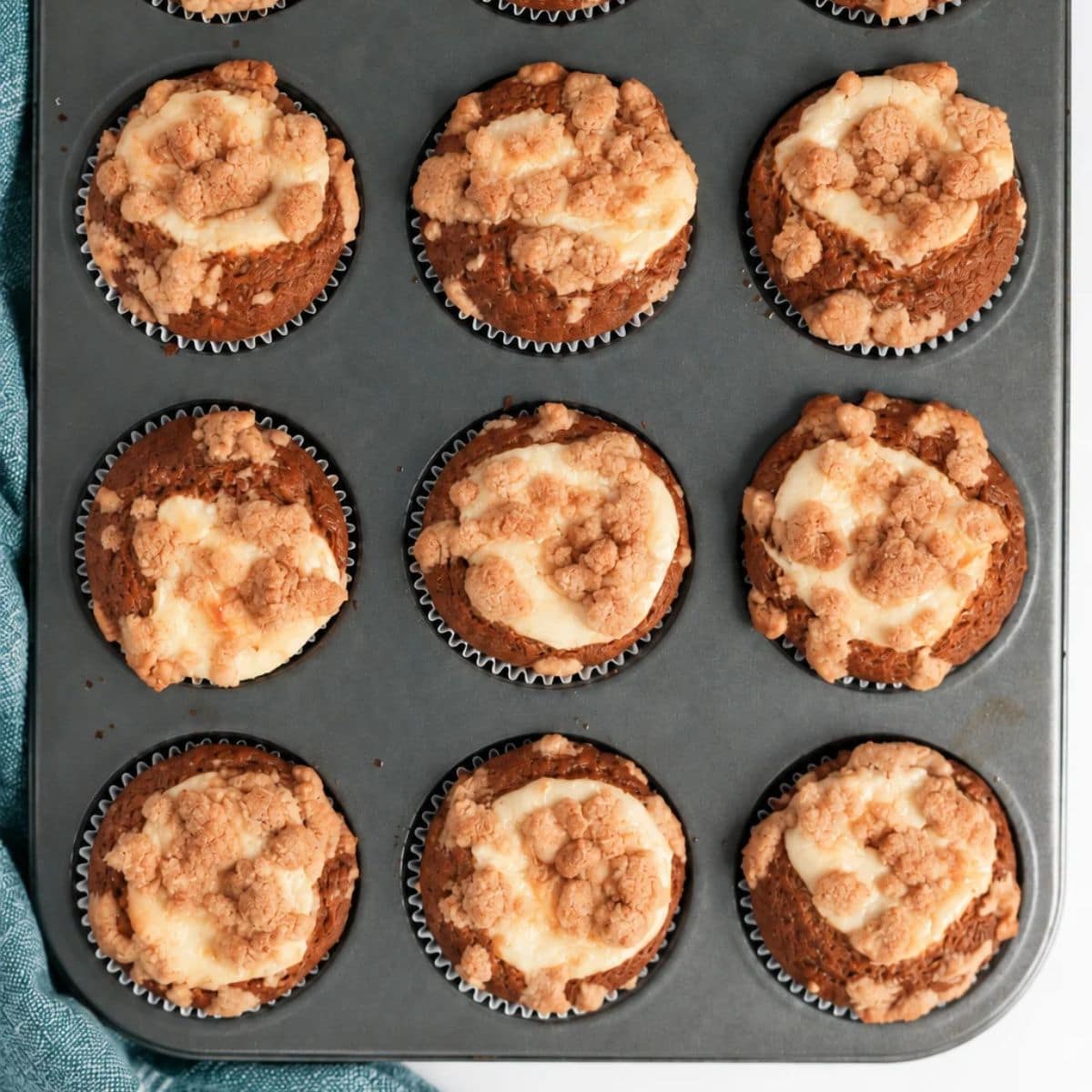 A muffin tin filled with twelve freshly baked Pumpkin Cream Cheese Muffins, each topped with a crumbly streusel and a luscious cream cheese swirl. A blue cloth is partially visible to the left.