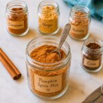 Five glass jars on a white surface, labeled with spices—Cinnamon, Ginger, Nutmeg, Clove, and Pumpkin Spice Mix—with a measuring spoon in the Pumpkin Spice Mix jar and two cinnamon sticks beside them.