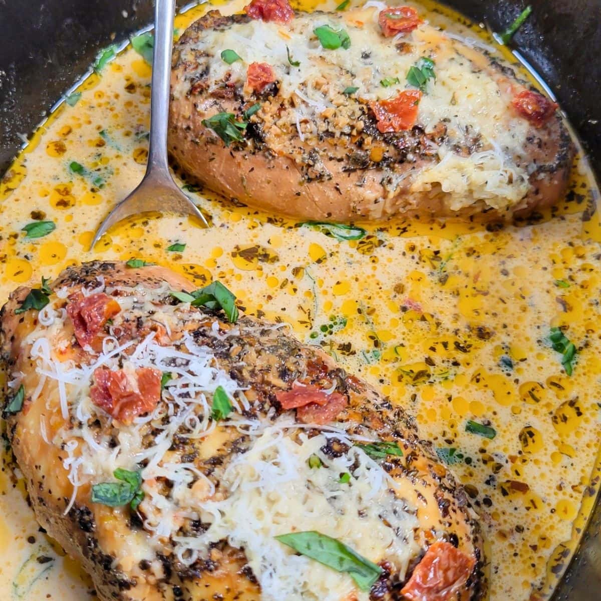 Two chicken breasts topped with herbs, cheese, and sun-dried tomatoes, simmering in a creamy sauce in a skillet.