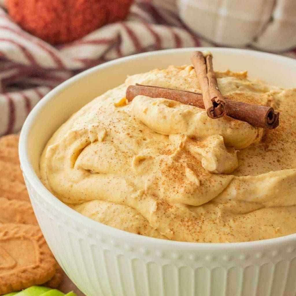 A bowl of creamy pumpkin and cream cheese dip topped with cinnamon sticks and a dusting of spice.
