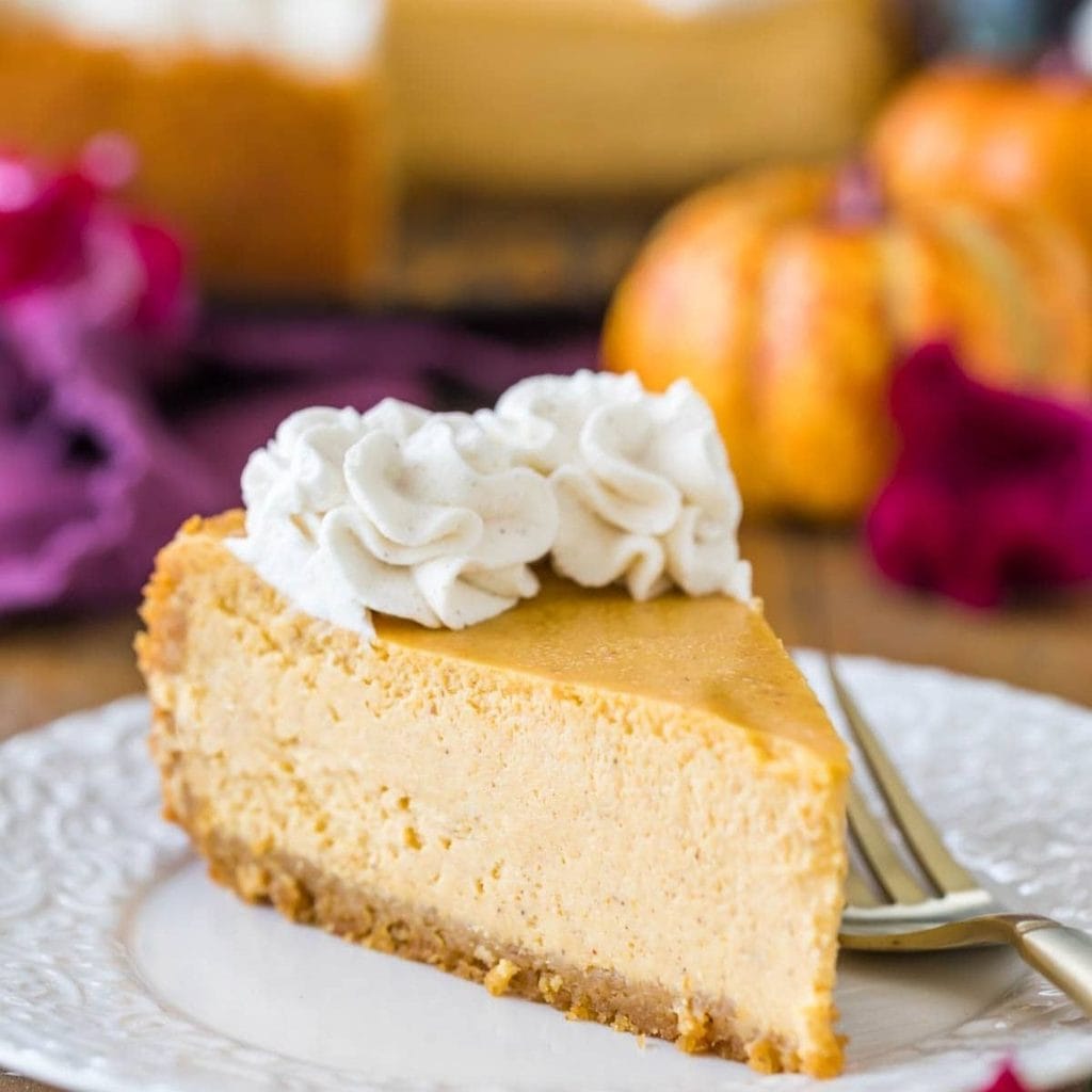 A slice of pumpkin and cream cheese recipe cheesecake topped with whipped cream on a decorative white plate with a fork.