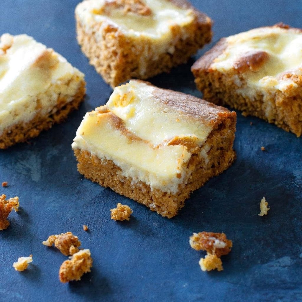 Four square pieces of pumpkin and cream cheese recipe cheesecake bars on a dark blue surface.